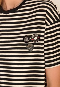 T-shirt rayé noir et blanc avec trois épingles : deux rondes avec du texte et une en forme de médiator avec un design graphique.