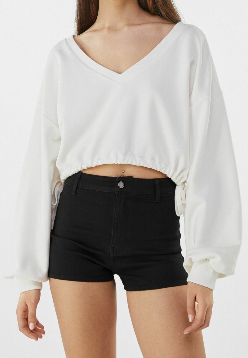 Femme portant un haut blanc court à manches longues et col en V et un short noir taille haute, montrant un piercing au nombril.