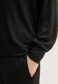 Schwarzes, strukturiertes Oberteil mit leichtem Schimmer, das lange Ärmel und eine lockere Passform bietet. Hand in der Tasche von schwarzen Hosen, die glattes Material zeigen.