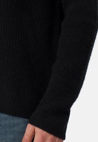 Svart ribbad stickad tröja med en mjuk textur, åtsittande ärmmuddar och en avslappnad passform, kombinerad med blå denim som syns nederst.