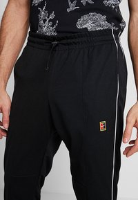 Zwarte joggers met een elastische tailleband en een trekkoord, voorzien van witte strepen aan de zijkanten en een klein gekleurd logo op het linkerbeen.