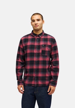 Jeune homme portant une chemise à carreaux rouges et noirs à manches longues avec des boutons et un jean bleu foncé, debout devant un fond uni.