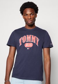 T-shirt de algodão azul-marinho com grande texto vermelho "TOMMY", "JEANS" em menor dimensão abaixo, e um logótipo no punho esquerdo. Decote em crew clássico.