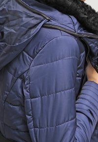 Chaqueta de plumas azul marino con textura acolchada, capucha forrada de piel y cierre de cremallera. El material parece suave e insulated, diseñado para proporcionar calor.