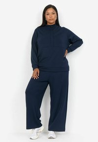 Tumedat sinised, pika varrukaga pullover kõrge kaelusega, mis on ühendatud sobivate laia säärega pükstega pehmest kangast. Ees on pool-zipper.
