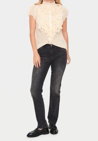 Donna che indossa una camicetta bianca a maniche corte con ruches e macchie gialle, jeans skinny neri sbiaditi e mocassini neri, in piedi su sfondo bianco.