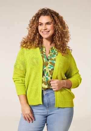 Gebreide cardigan in fel limoen groen, met ribbels aan de mouwen en onderrand, gedragen over een gebloemde groen en bruine blouse met een knoopsluiting bij de hals.