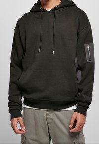 Sudadera negra con capucha, cordones en el frente y un bolsillo con cremallera en la manga izquierda, usada sobre una camisa blanca y pantalones cargo grises.