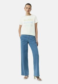 Witte korte mouwen t-shirt met multikleurige "BLOOM" tekst, gecombineerd met lichtblauwe wijde denim jeans en lichtgekleurde blokhak sandalen.