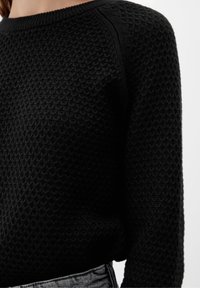 Pull noir texturé à manches longues porté par une personne, visible du cou à la taille, associé à un jean en denim bleu clair.