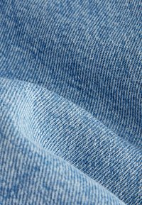 Tissu en denim aux tons bleu clair, présentant une texture lisse avec un motif en chevron subtil et des détails de couture visibles.