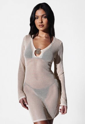 Jeune femme portant une robe beige en maille ajourée à manches longues sur un bikini noir, debout devant un fond clair uni.