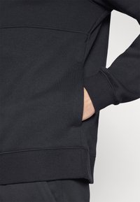 Sudadera negra con una textura suave, con cuello redondo, puños acanalados y un bolsillo lateral. El diseño incluye costuras mínimas y un corte holgado.