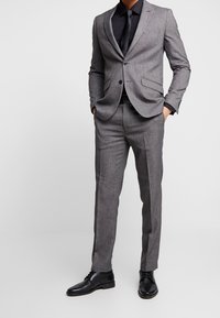 Man i en skräddarsydd grå kostym med svart skjorta, svart slip, svart bälte och svarta läderskor, stående med händerna i fickorna.