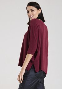 Burgunderfarbener Strickpullover mit Dreiviertelärmeln, lässiger Schnitt und seitlichen Schlitzen. Kombiniert mit schwarzen, eng geschnittenen Hosen, glatte Textur.