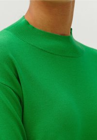 Gros plan sur une personne portant un pull col montant côtelé vert vif en tissu finement tricoté.