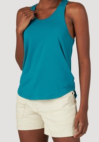 Kvinna i turkos ärmlös topp och beiga shorts, med handen vilande nära kragen, mot en enfärgad ljus bakgrund.