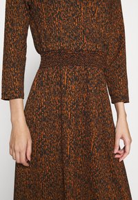 Nahaufnahme von Händen und Taille, die ein langärmeliges, orangefarbenes und schwarzes gemustertes Kleid mit einem gerafften, strukturierten Bund tragen.