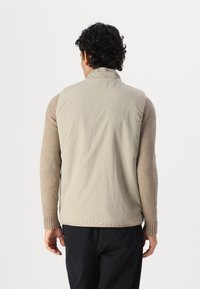 Leichte Weste in Beige mit einem glatten, gesteppten Rücken und flauschigen, beigen Ärmeln. Sie verfügt über einen Stehkragen und gerippte Bündchen.