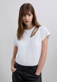 White short-sleeve top with an asymmetrical design, featuring a decorative gold chain on the shoulder and a soft, smooth texture.