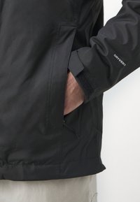 Chaqueta impermeable negra con una textura suave, que cuenta con un bolsillo lateral con cremallera y detalles en Velcro en los puños. Marca "DRYVENT" en la manga.