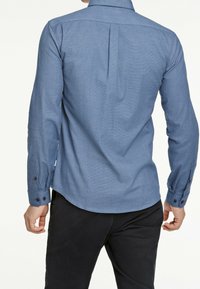 Camicia blu con tessuto strutturato, maniche lunghe, dettaglio con piega sul retro e polsini con bottoni. Abbinata a pantaloni scuri.