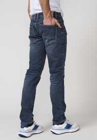 Jeans de mezclilla azul oscuro con corte ajustado, detalles de costura visibles y dos bolsillos traseros. Llevados con zapatillas blancas con acentos azules.