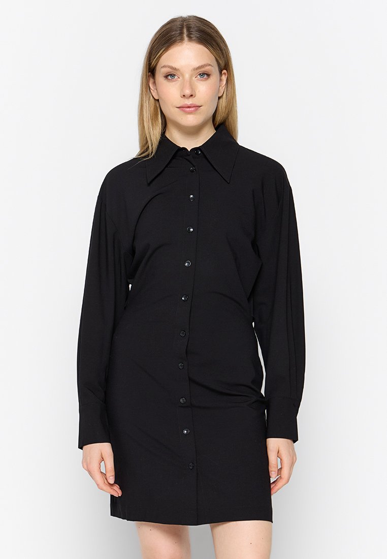 Frau in einem schwarzen, langärmligen Kleid mit Knopfleiste und weitem Kragen, vor einem einfachen weißen Hintergrund stehend.