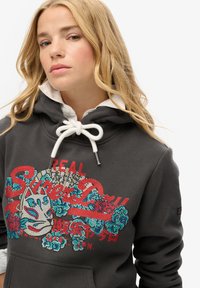 Sudadera gris con cordón y acentos blancos, que presenta adornos de strass coloridos y gráficos de texto en el frente.