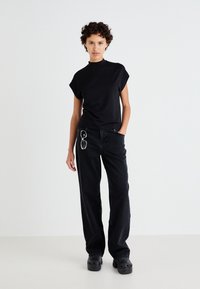 Ženska s kratkimi kodrastimi lasmi, oblečena v črno majico s visokim ovratnikom, črne široke hlače in črne platforme, stoji ob belem zidu, z sončnimi očali pripetimi na oblačila.