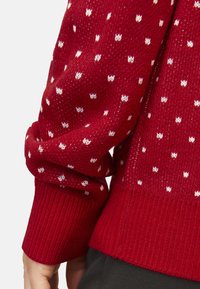 Czerwony sweter z dzianiny z białymi kropeczkami, ściągaczami przy rękawach i dolnym hemie. Materiał ma miękką teksturę i luźny krój.