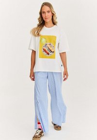 Bílý tričko s krátkým rukávem se vzorem pestrého jídla na talíři, spárované s modro-bíle pruhovanými širokými kalhotami.