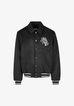 Melna varsity stila jaka ar snap pogām, balta emblēma kreisajā krūtī un melnbaltām svītrām pie piedurknēm un jostas.
