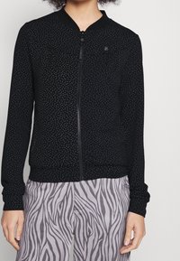 Svart zip-up bomberjacka med små vita prickar, ribbat krage och ärmslut, samt en figurnära nederkant. I kombination med ljusgrå zebramönstrade byxor.