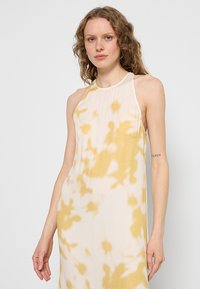 Żółto-biała sukienka na szyję z motywem tie-dye, wykonana z lekkiego, lejącego się materiału. Charakteryzuje się minimalistycznym wzornictwem i odkrytymi plecami.