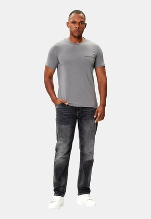T-shirt grigio a maniche corte con un logo discreto, abbinato a jeans in denim scuro e sneakers bianche, caratterizzato da un design semplice.
