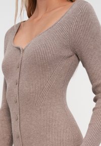 Ριγέ πλεκτό ολόσωμο σε ανοιχτό καφέ χρώμα με V-λαιμόκοψη, κλείσιμο με κουμπί και μακριά μανίκια, που διαθέτει εφαρμοστή σιλουέτα και υφασμάτινη υφή.