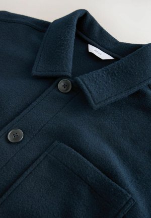 Tamsiai mėlynas vilnos mišinio marškiniai striukė su tekstūruotu paviršiumi, turinti klasikinį apykaklę, dvi juodas mygtukus ir krūtinės kišenę.