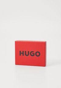 Rød rektangulær kasse med en glat overflade, der har ordet "HUGO" i fed sort skrifttype på den ene side.