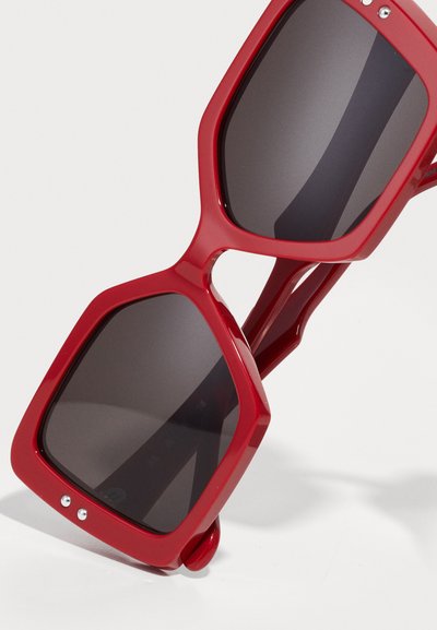 Occhiali da sole rossi oversize con una montatura geometrica, lenti scure e materiale plastico lucido. Caratteristiche includono dettagli metallici e aste spesse.