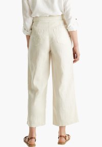 Personne portant un pantalon crème taille haute à jambes larges avec poches arrière, une chemise blanche à manches retroussées et des sandales marron à lanières, debout de dos.