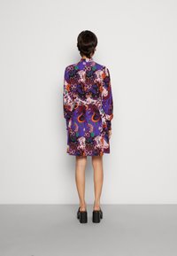 Kurzes, lila Blumenmusterkleid mit langen Ärmeln, tailliert und mit lebhaften Mustern. Kombiniert mit klobigen schwarzen High Heels.