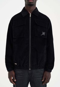 Mustan corduroy-takin, jossa on vetoketjukiinnitys, kaksi rintataskua ja elastinen helma. Vasemmassa rinnassa brodeerattu logo.