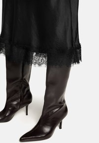 Schwarze, spitz zulaufende Lederstiefel mit Absatz, schlankem Design und seitlichem Reißverschluss, getragen zu einem schwarzen Kleid mit Spitzenabschluss.