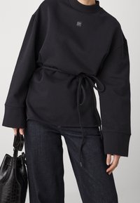 Persona che indossa una maglietta nera a maniche lunghe con un nodo in vita, jeans scuri e tiene in mano una borsa a tracolla in pelle nera con texture.