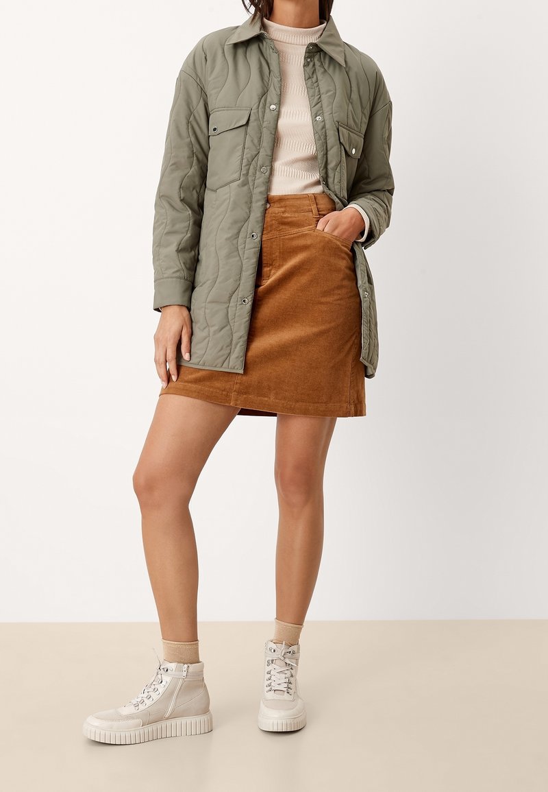Veste matelassée verte avec deux poches avant, haut à manches longues beige à côtes, jupe en velours côtelé marron et baskets montantes de couleur claire.