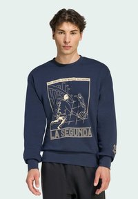 Sudadera azul marino con un gráfico en crema que representa una escena de fútbol y el texto "LA SEGUNDA", con puños y dobladillo acanalados.