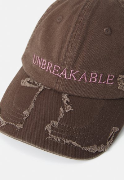 Brūna nolietota beisbola cepure ar rozā izšūtu tekstu "UNBREAKABLE" uz priekšējā paneļa, rādīta auduma nolietojums un bārkstis.