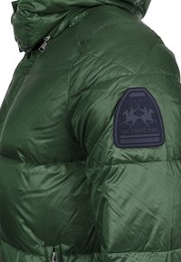 Green padded jacket with a shiny finish, featuring a navy logo patch on the shoulder, zip fastener, and quilted texture.