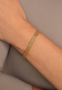 Bracciale color oro con una design piatto e intrecciato, caratterizzato da una finitura testurizzata, poggiato su un polso su uno sfondo di tessuto neutro.